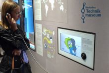 Foto: Frau steht vor einer Medienstation und schaut ein Erklärvideo - Logo Deutsches Technikmuseum
